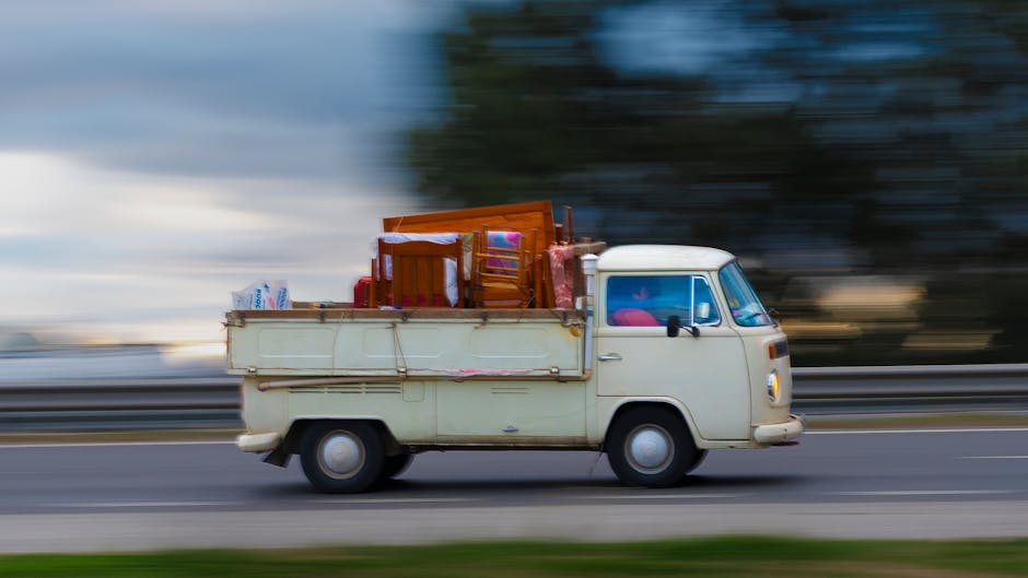 A vintage cream-colored transporter van with a compact, rounded shape is driving along a motorway, loaded with various household items for a home relocation. Inside the vehicle's open cargo area, wood