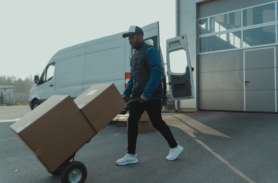 A man wearing a black cap, blue jacket, and black trousers is captured during a home relocation process, pushing a hand truck loaded with large cardboard boxes wrapped in plastic. He is walking on a p