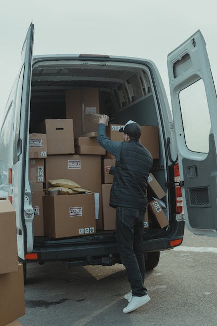 A man wearing a black cap, blue jacket, and black trousers is captured during a home relocation process, pushing a hand truck loaded with large cardboard boxes wrapped in plastic. He is walking on a p