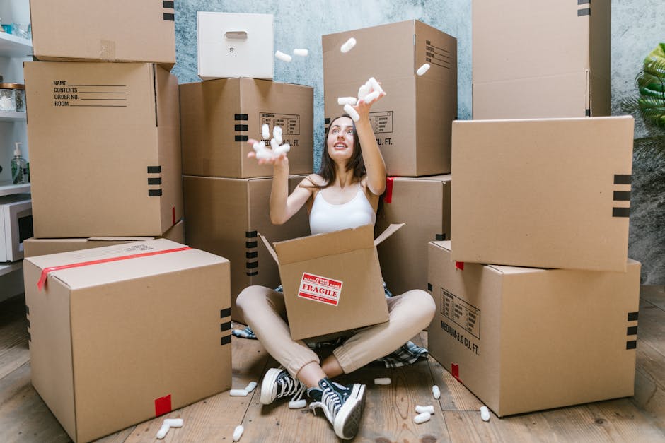 A young woman with long dark hair, wearing a white tank top and beige pants, is sitting cross-legged on a wooden floor inside a home, surrounded by stacked cardboard moving boxes of various sizes, som