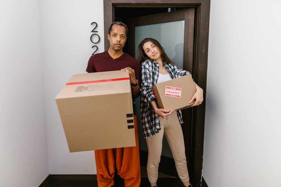 A young woman with long dark hair, wearing a white tank top and beige pants, is sitting cross-legged on a wooden floor inside a home, surrounded by stacked cardboard moving boxes of various sizes, som