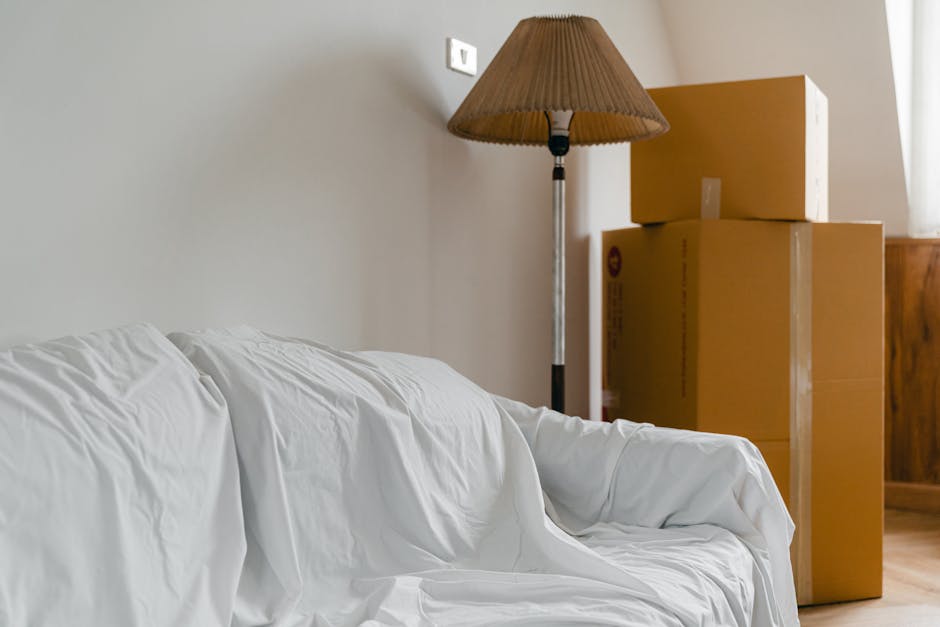 A woman with curly dark hair, dressed in a green t-shirt and blue jeans, is preparing a sofa for a house removal. She is wrapping the sofa in protective white plastic sheeting to safeguard the furnitu
