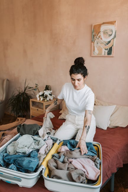 A young woman with shoulder-length brown hair, wearing a light blue long-sleeve shirt, is sitting at a wooden table inside a home, sealing a large black plastic bag containing clothing or textiles wit