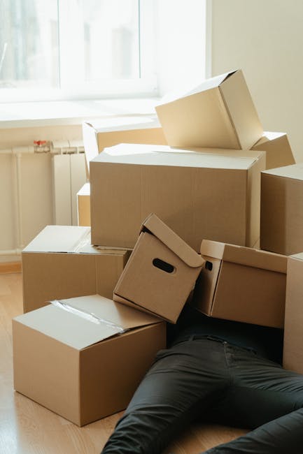 Close-up view of three cardboard boxes used for home relocation or packing, with white labels indicating contents; the top box is labeled 'SUMMER CLOTHS' and positioned on a wooden floor inside a prop